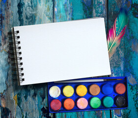 Top view close-up photo of sketchbook, palette, watercolor, colored feather, rustic wood background. Copy space for text. Natural, boho, ethnic or hipster vintage mock up.