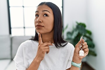 Young hispanic woman holding keys of new home thinking concentrated about doubt with finger on chin...