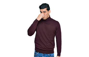 Fototapeta premium Handsome hispanic man wearing casual turtleneck sweater smelling something stinky and disgusting, intolerable smell, holding breath with fingers on nose. bad smell