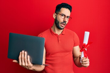 Young hispanic man holding laptop and diploma clueless and confused expression. doubt concept.