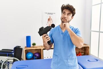 Hispanic physiotherapy man working at pain recovery clinic with muscle gun serious face thinking about question with hand on chin, thoughtful about confusing idea