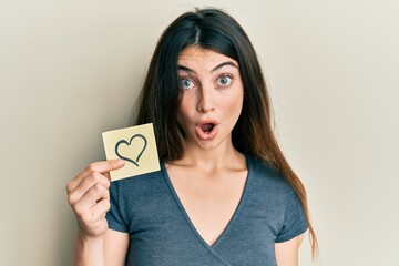 Young caucasian woman holding heart reminder scared and amazed with open mouth for surprise,...