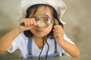 Little kid girl asian wearing a white hat and jeans jumpsuit and xploring nature with a magnifying glass. Which increases the development and enhances outside the classroom learning skills concept.