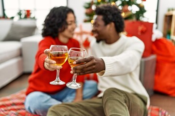 Young interracial couple smiling happy and toasting with champagne sitting on the floor by christmas tree at home.