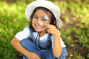Little kid girl asian wearing a white hat and jeans jumpsuit and xploring nature with a magnifying...