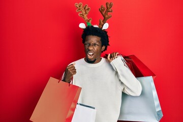Young african american man wearing deer christmas hat holding shopping bags angry and mad screaming frustrated and furious, shouting with anger. rage and aggressive concept.