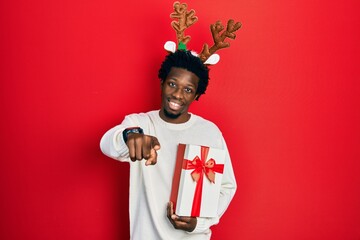 Young african american man wearing deer christmas hat holding gift pointing to you and the camera with fingers, smiling positive and cheerful
