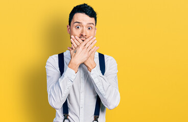 Hispanic man with beard wearing hipster look with bow tie and suspenders shocked covering mouth with hands for mistake. secret concept.