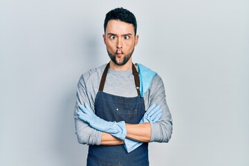 Young hispanic man wearing apron with arms crossed making fish face with mouth and squinting eyes, crazy and comical.