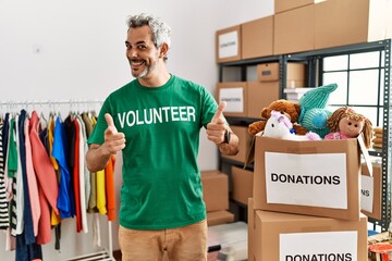 Middle age hispanic man wearing volunteer t shirt at donations stand pointing fingers to camera with happy and funny face. good energy and vibes.