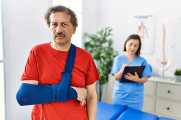 Southeast asian man wearing wearing arm on sling at rehabilitation clinic thinking attitude and sober expression looking self confident