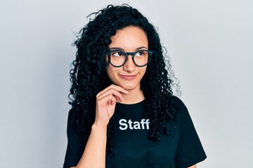 Young hispanic woman with curly hair wearing staff t shirt with hand on chin thinking about...