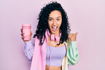 Young hispanic woman with curly hair wearing sportswear and headphones drinking cup of coffee pointing thumb up to the side smiling happy with open mouth