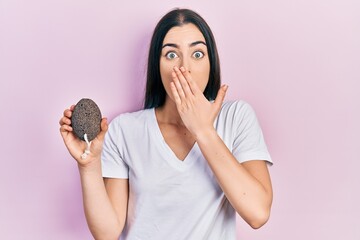 Beautiful woman with blue eyes holding pumice stone covering mouth with hand, shocked and afraid...