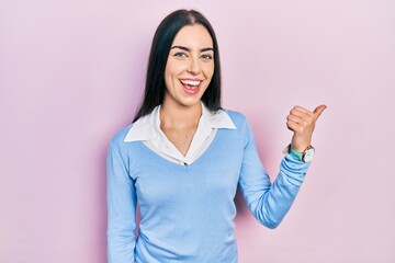 Beautiful woman with blue eyes standing over pink background smiling with happy face looking and pointing to the side with thumb up.