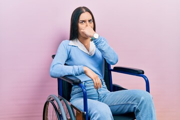 Fototapeta na wymiar Beautiful woman with blue eyes sitting on wheelchair smelling something stinky and disgusting, intolerable smell, holding breath with fingers on nose. bad smell