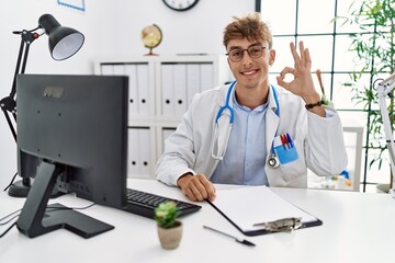 Young caucasian doctor man working at the clinic smiling positive doing ok sign with hand and fingers. successful expression.