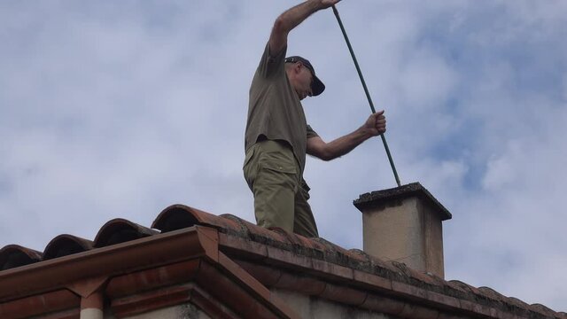 The man on the roof cleans the chimney with long brush for the fireplace.
