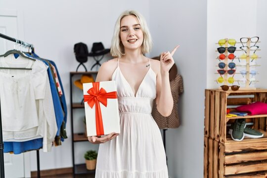 Young caucasian woman holding gift at retail shop smiling happy pointing with hand and finger to the side