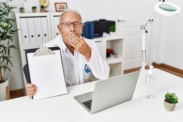 Senior doctor man holding clipboard with blank space covering mouth with hand, shocked and afraid for mistake. surprised expression