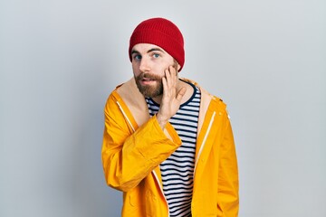 Caucasian man with beard wearing yellow raincoat hand on mouth telling secret rumor, whispering malicious talk conversation