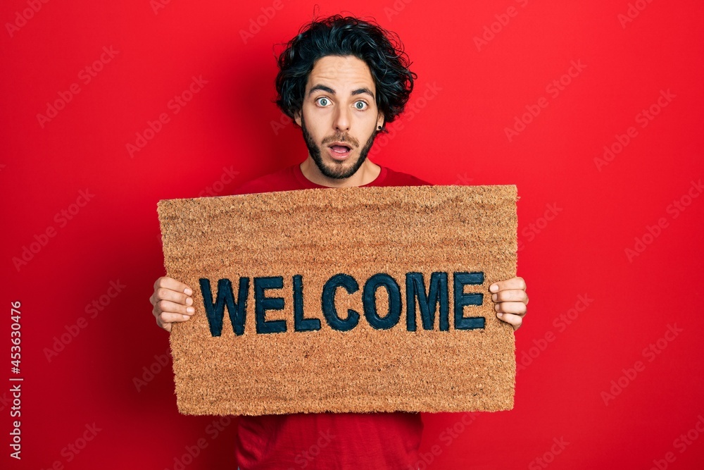 Sticker handsome hispanic man holding welcome doormat afraid and shocked with surprise and amazed expression