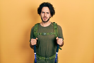 Handsome hispanic man wearing backpack holding trekking poles clueless and confused expression....