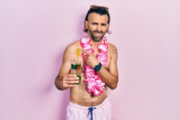 Young hispanic man wearing swimsuit and hawaiian lei drinking tropical cocktail pointing aside...