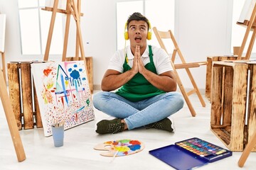 Young hispanic man at art studio begging and praying with hands together with hope expression on...