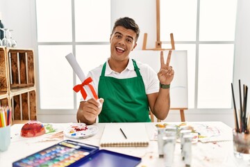 Young hispanic man at art studio holding degree smiling with happy face winking at the camera doing victory sign with fingers. number two.