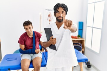 Fototapeta premium Young hispanic man working at pain recovery clinic with a man with broken arm pointing with finger up and angry expression, showing no gesture