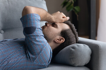Tired frustrated millennial man lying on sofa at home, thinking over problems, feeling stress,...