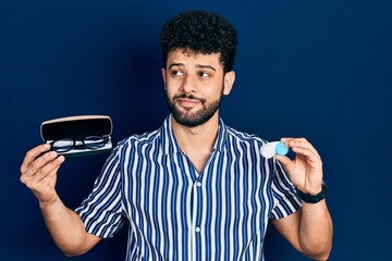 Young arab man with beard holding glasses and contact lenses smiling looking to the side and...