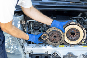 Automotive technician holding used car pressure plate, clutch disc and release bearing in front of the vehicle engine - obrazy, fototapety, plakaty