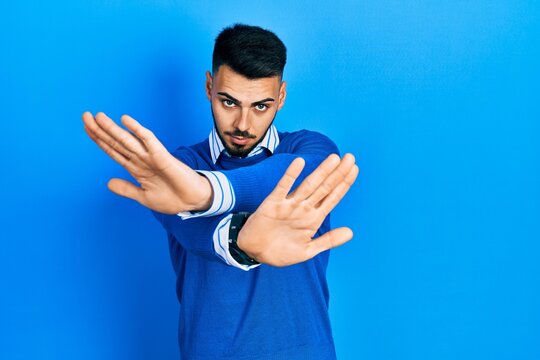 Young hispanic man with beard wearing casual blue sweater rejection expression crossing arms and palms doing negative sign, angry face