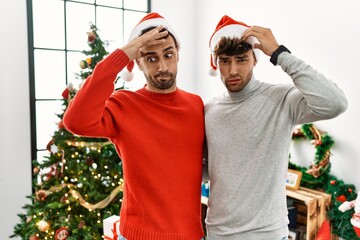 Young gay couple standing by christmas tree wearing hat worried and stressed about a problem with hand on forehead, nervous and anxious for crisis