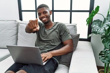 Young african american man using laptop at home sitting on the sofa pointing to you and the camera with fingers, smiling positive and cheerful