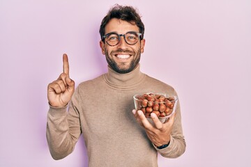 Handsome man with beard holding bowl of chestnuts smiling with an idea or question pointing finger with happy face, number one