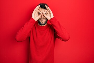Handsome man with beard wearing casual red sweater doing ok gesture like binoculars sticking tongue out, eyes looking through fingers. crazy expression.
