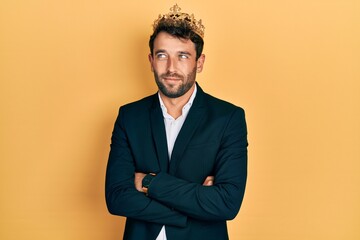 Handsome man with beard wearing business suit with arms crossed gesture and king crown smiling looking to the side and staring away thinking.