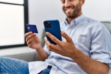 Young hispanic man using smartphone and credit card sitting on the sofa at home.