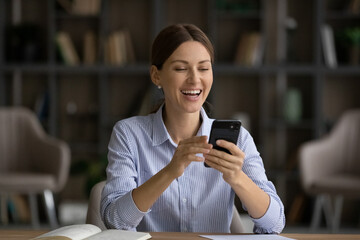 Excited laughing businesswoman looking at phone screen, reading good news, sitting at work desk,...