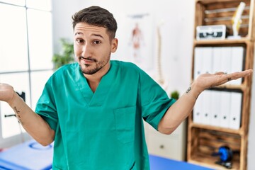 Young physiotherapist man working at pain recovery clinic clueless and confused expression with arms and hands raised. doubt concept.