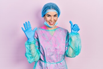 Young brunette woman wearing surgeon uniform and medical mask showing and pointing up with fingers number six while smiling confident and happy.