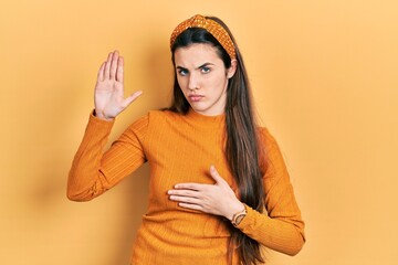 Young brunette teenager wearing casual yellow sweater swearing with hand on chest and open palm,...