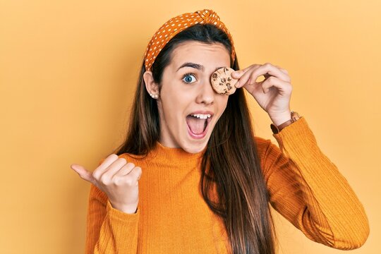 Young brunette teenager holding cookie over eye pointing thumb up to the side smiling happy with open mouth