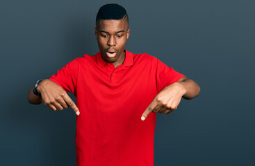 Fototapeta premium Young african american man wearing casual red t shirt pointing down with fingers showing advertisement, surprised face and open mouth