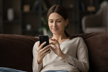 Close up satisfied young woman using smartphone, sitting on couch at home, pleasant young female...
