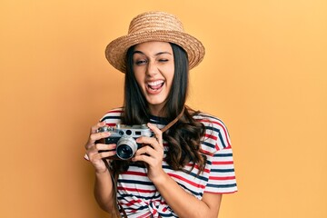 Young hispanic woman wearing summer hat holding vintage camera winking looking at the camera with...