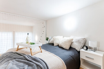 Fototapeta na wymiar Simple bedroom in loft apartment in wooden style with double bed, two nightstands with lamps and two glasses of wine with bottle on dining tray.Bedroom is lit by circular bright light mounted on wall.